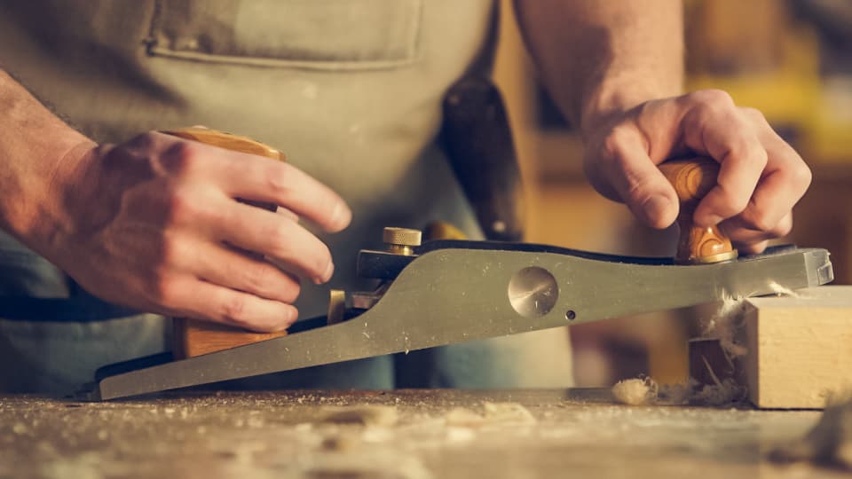 A carpenter's hands running a hand plane along a board, shavings curling off it